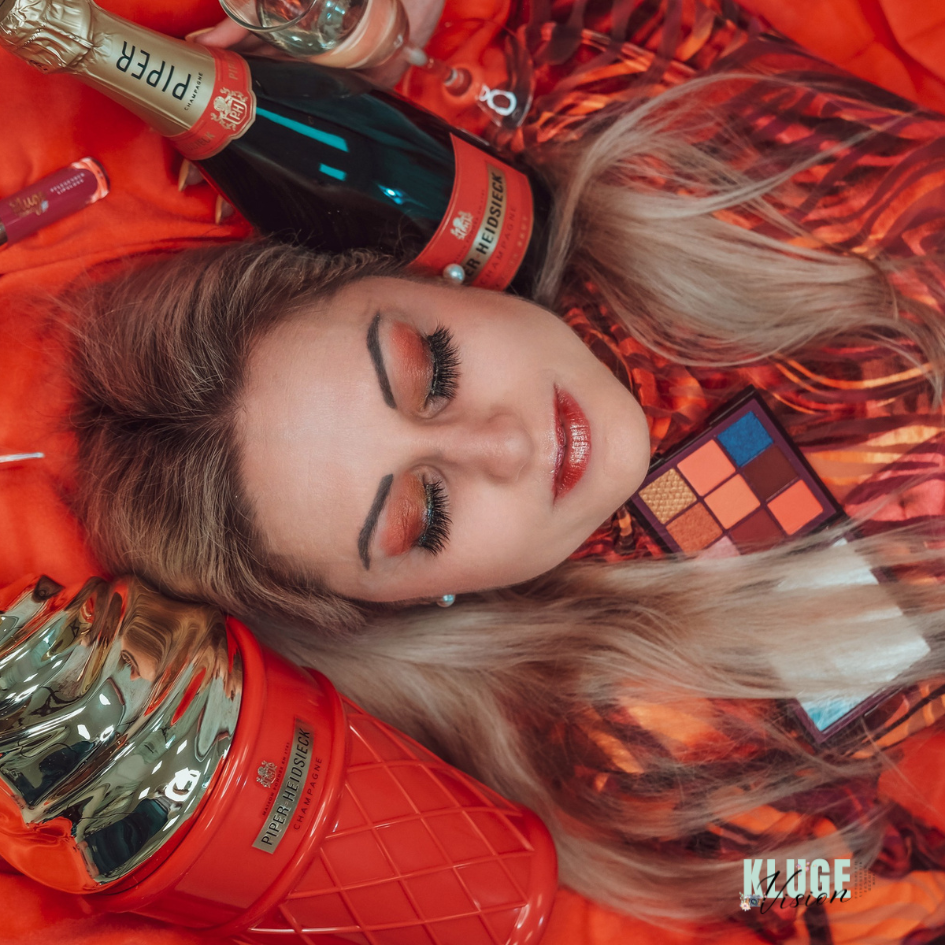 Stilvolle Frau mit orangefarbenem Augen-Make-up, einer roten Palette und Champagnerflasche, dargestellt in einem luxuriösen Arrangement.