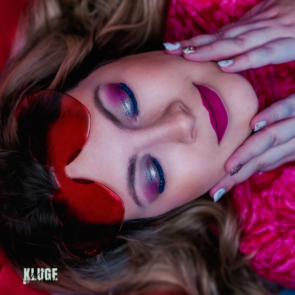 Kreatives Porträt einer Frau mit pinkem Augen-Make-up, knallpinkem Lippenstift und einer herzförmigen Brille, vor einem lebhaften Hintergrund.