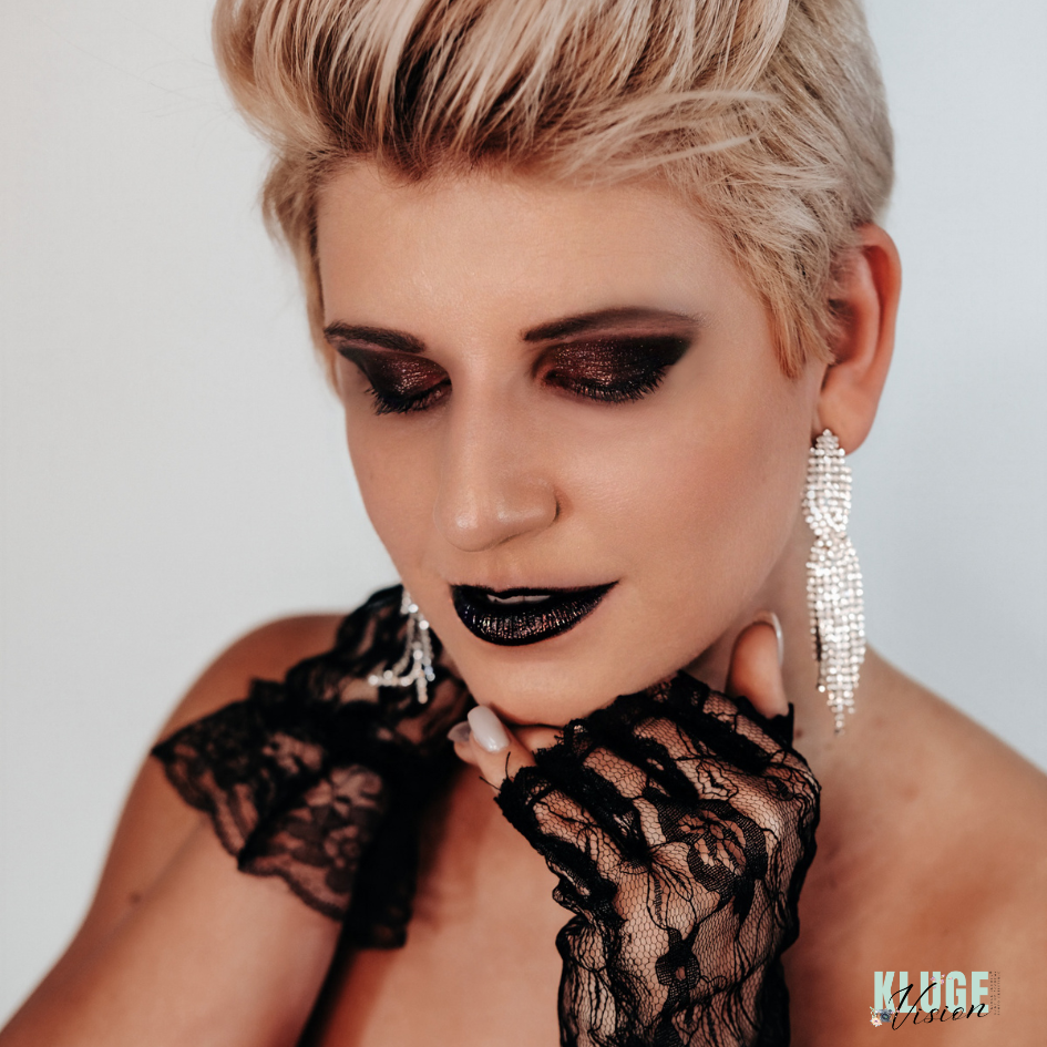 Porträt einer Frau mit intensivem, schwarzem Augen-Make-up und Lippenstift, ergänzt durch elegante, funkelnde Ohrringe und schwarze Spitzenhandschuhe.